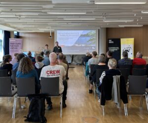 Bild einer Versammlung bei der einige Menschen von hinten zu sehen sind und auf zwei Vortragende mit Präsentationsbildschirm schauen auf dem "Initiativenwerkstatt Hamburg Repariert" steht